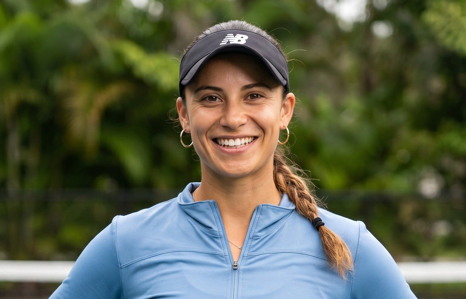 pro pickleball player lauren-hidalgo Smith in blue athletic jacket and cap standing on a tennis court with greenery in the background